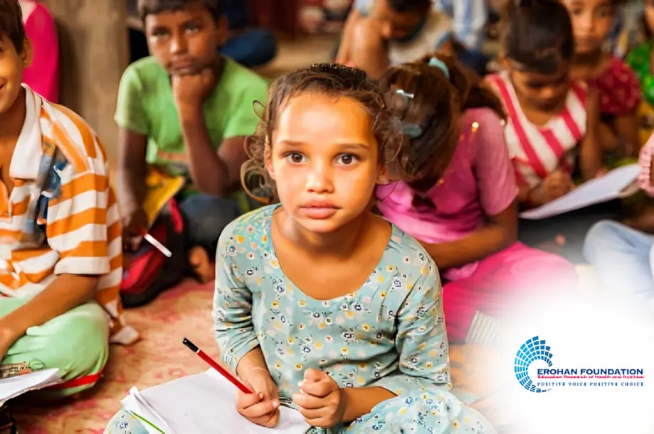 A group of children sitting on the floor, with one girl in focus holding a pencil and looking at the camera. The logo of EROHAN Foundation is visible in the bottom right corner.